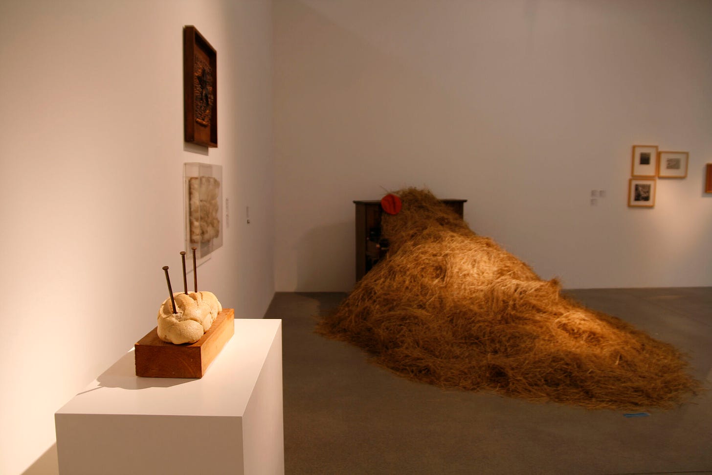 Lit bread installation in a room with framed paintings, next to installation of large haystack spilling out of piano
