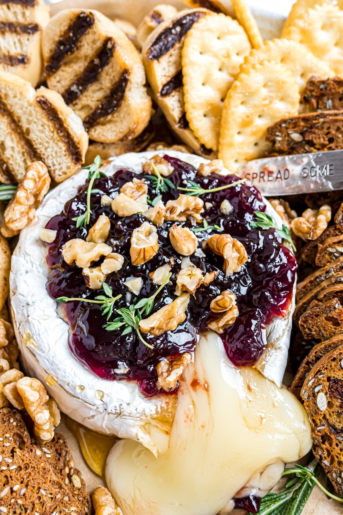 A wheel of baked brie topped with raspberry jam, walnuts, and fresh thyme, that has been cut open and there is melted cheese flowing onto the serving platter.