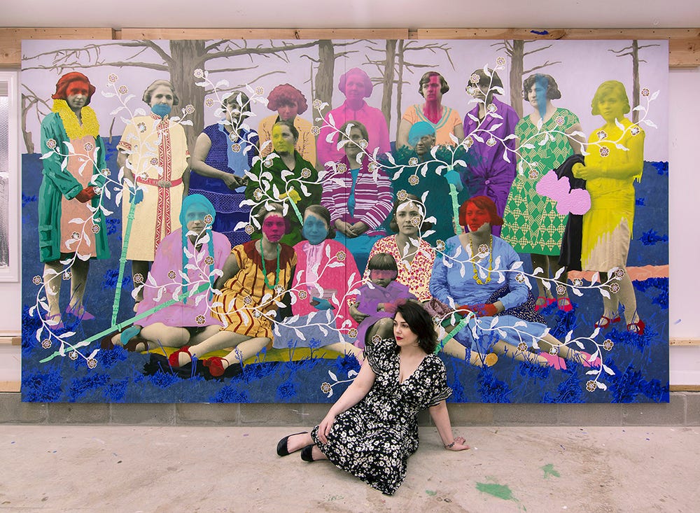 Daisy sits in front of a large painted photograph in her studio. Daisy is in a floral dress and has dark shoulder length hair. The large painting behind Daisy is a group of Bulgarian women sitting and standing together, with a photograph as its substrate. They are adorned with painted flowers, bright washes of color, blue grass, and a lavender sky.