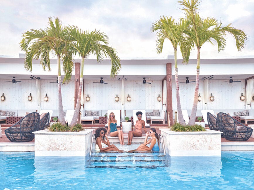 Four people relax by a pool with palm trees and lounge chairs in the background, sitting on submerged steps at a resort.