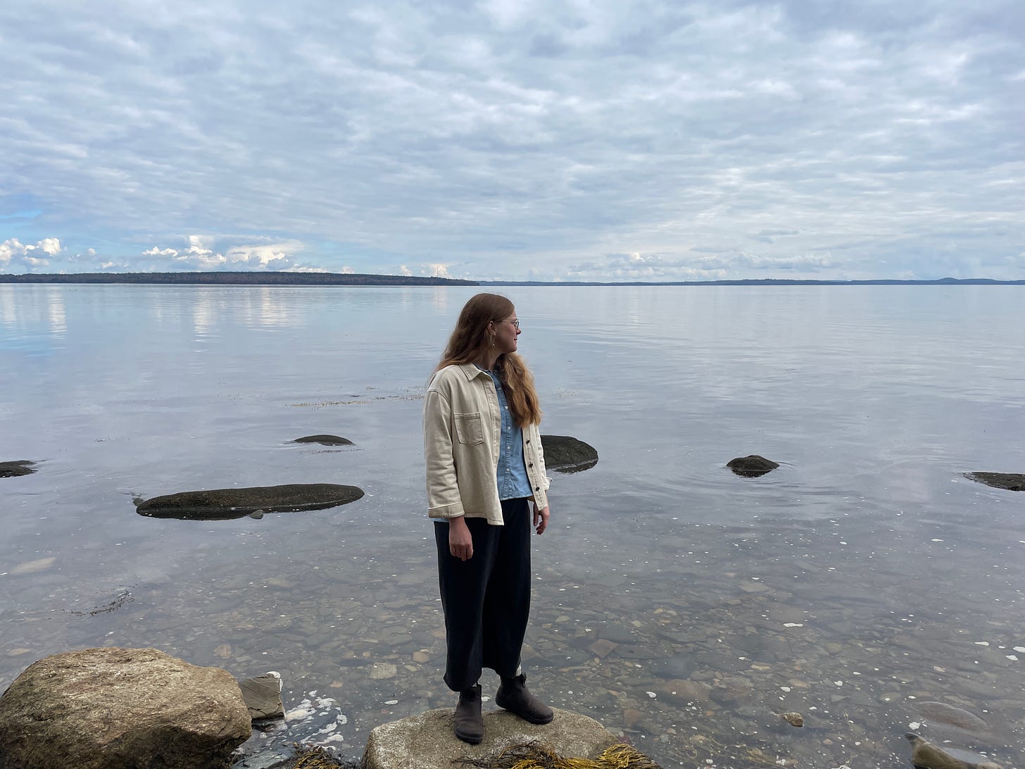 A women at center, wearing a cream jacket, denim shirt, dark grey pants and boots. She is also wearing thin, wire framed glasses and freshwater pearl earrings. She has long, strawberry blonde hair. She is standing on a rock at the edge of a river/bay. You can see clouds and the land on the horizon behind her. 