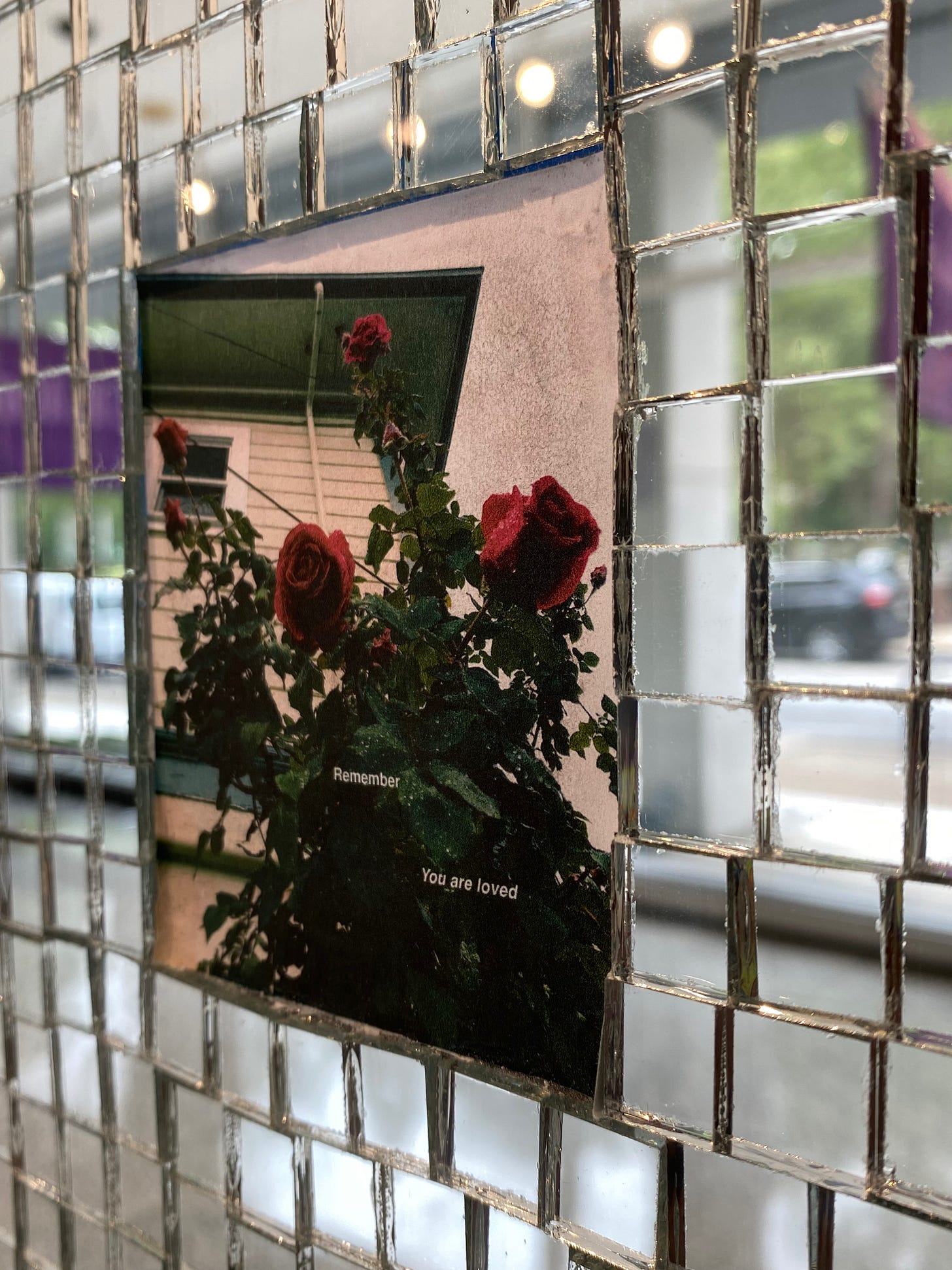 diagonal view of mirrored surface artwork with a photographic inset with a rose bush and house in background. White text reads: "Remember, you are loved"