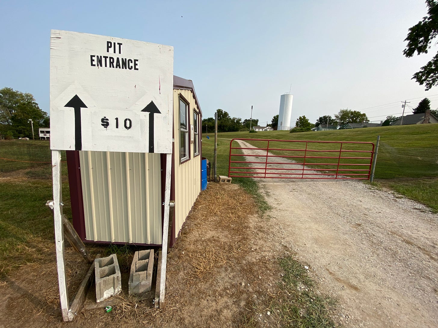 A photo of a sign along a gravel road. The sign says "Pit Entrance $10." The road is barred with a farm-style gate. Beyond it is grass, and a silo in the distance. 
