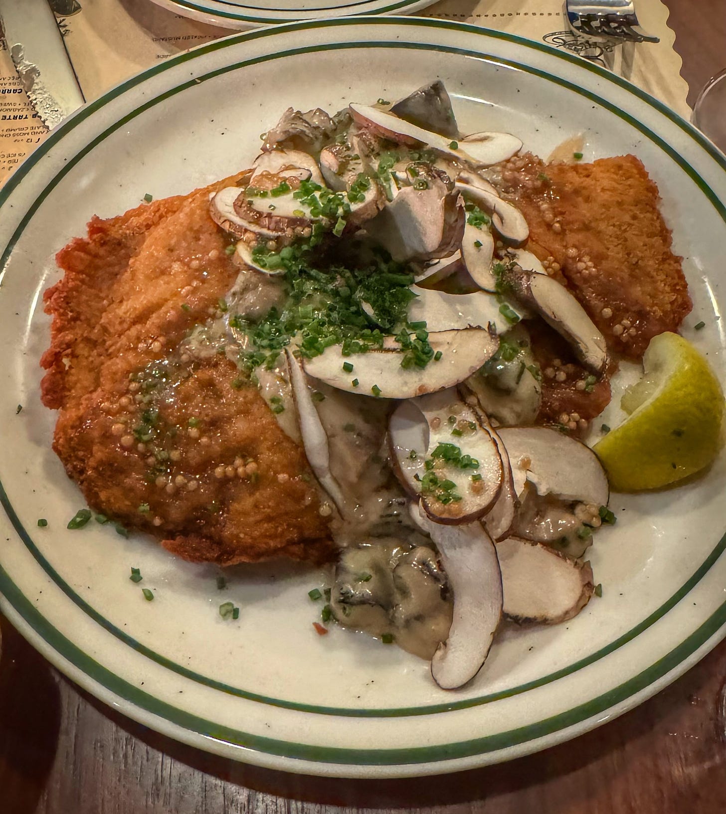 A large plate covered with a weinerschnitzel covered in sauce and large pieces of sliced mushroom and fermented mustard seeds. There is an already-squeezed wedge of lemon on the side of the plate. 