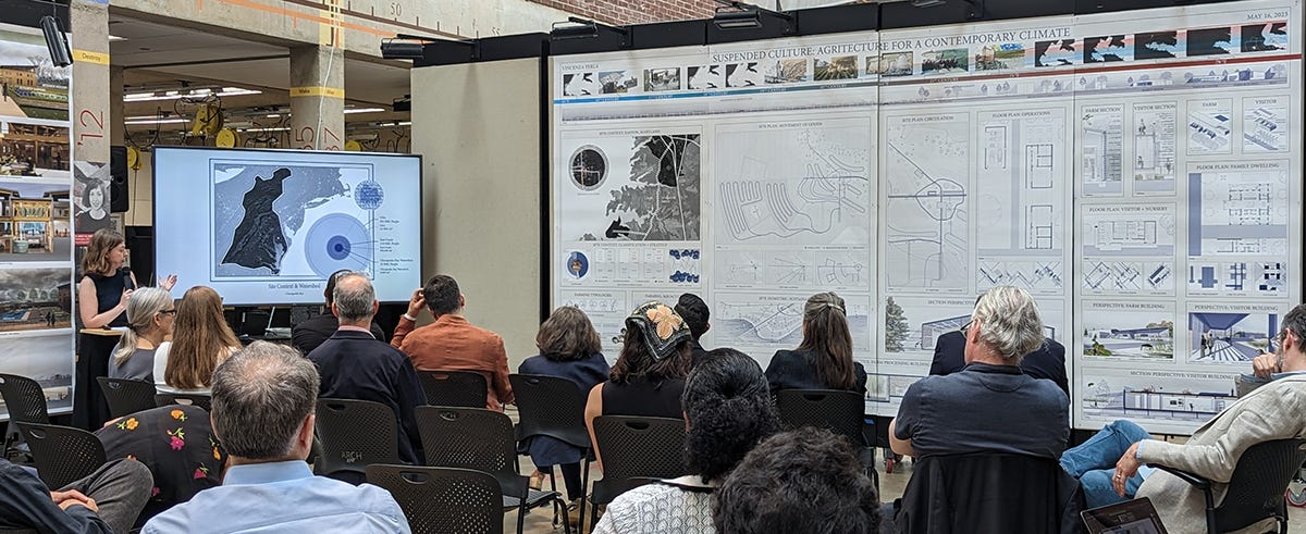 A master's thesis student presenting her project to an audience. Big drawings cover one wall and slides show on a large TV screen