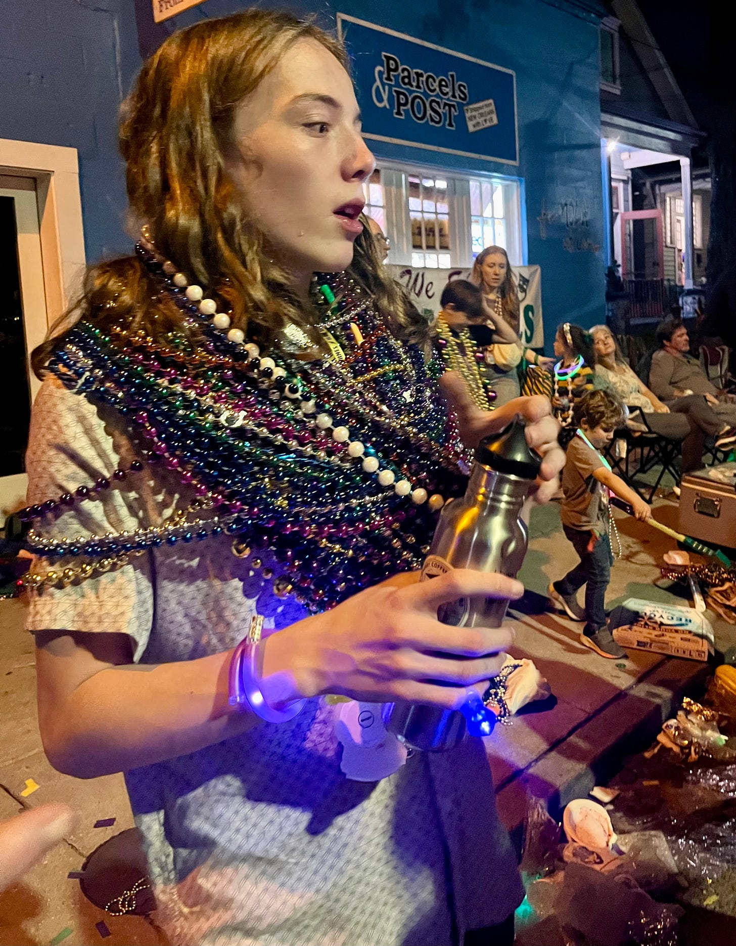 A teenager at a Mardi Gras parade wearing what looks to be a hundred strings of Mardi Gras beads around his neck, and flashing jewelry on his wrist. Other revelers sit, talk, and play in the background.