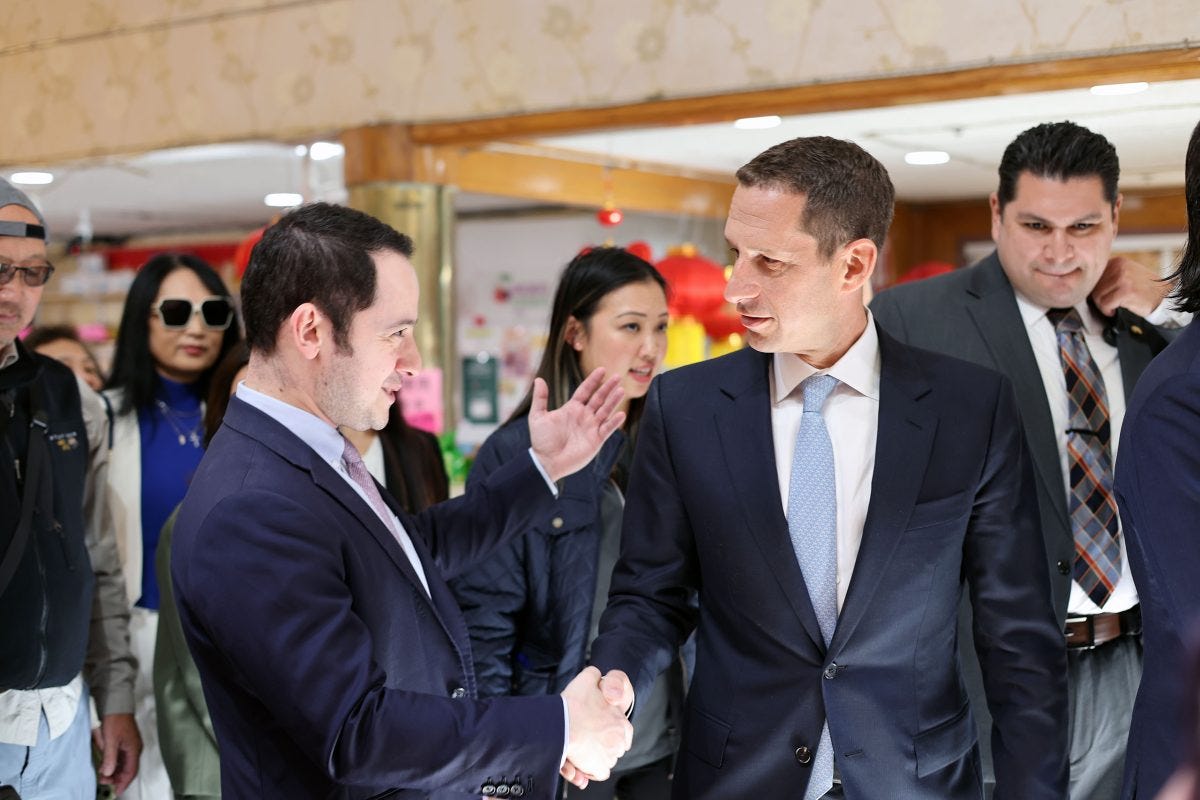 Two men in suits shake hands in a crowded indoor setting, surrounded by several people.