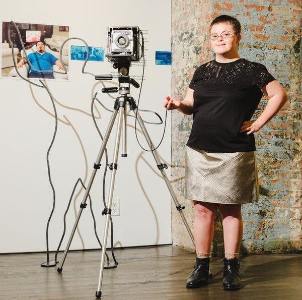 Rachel Handlin, wearing a black lace top and a silver skirt, stands beside her 4x5 film camera, which is on a tripod.