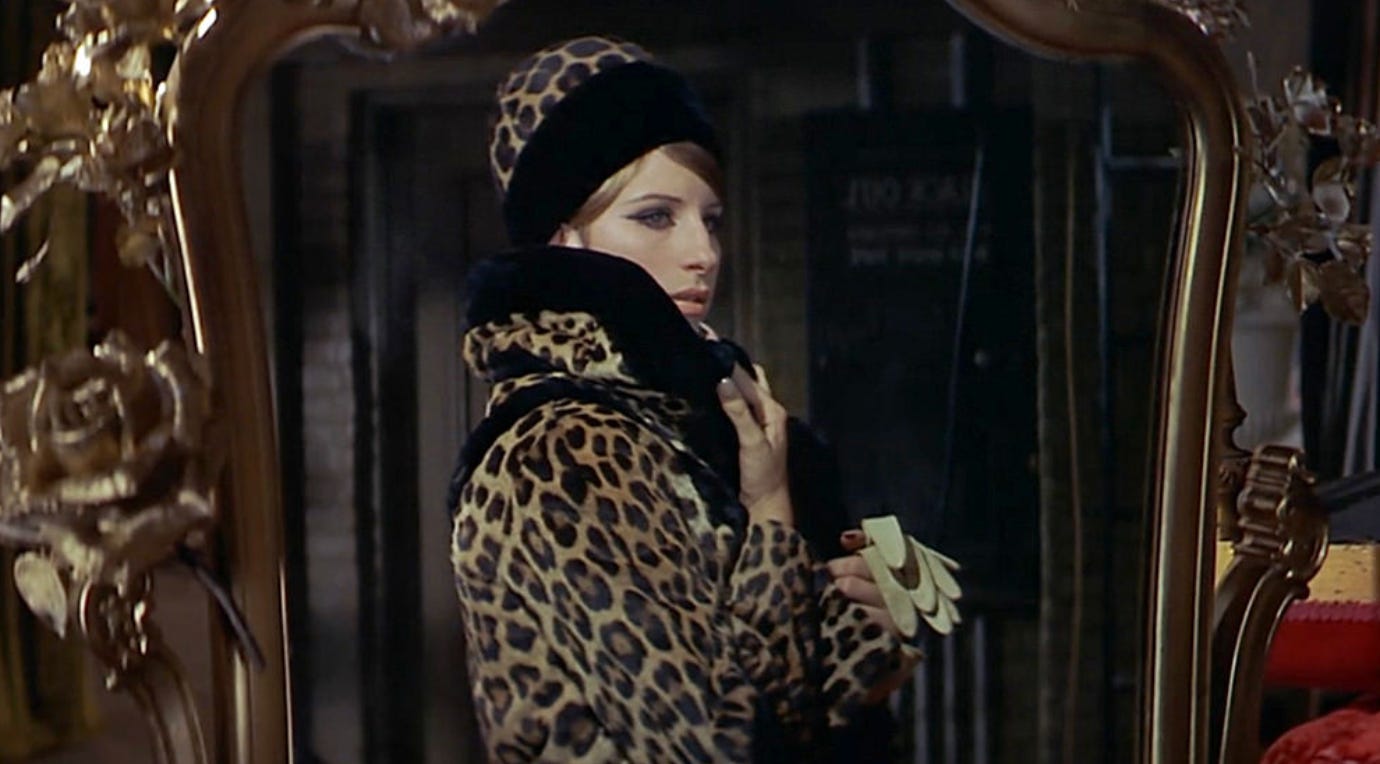 A screenshot from Funny Girl showing Barbra Streisand looking at herself in the mirror, while wearing a striking leopard-print coat and hat