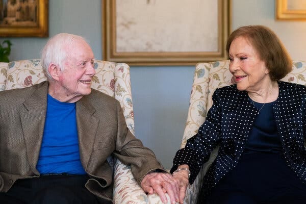 Mr. Carter, wearing a dark sports coat and a blue T-shirt, and Rosalynn Carter, wearing a dark jacket with small white dots and dark blouse, hold hands while seated in separate chairs.