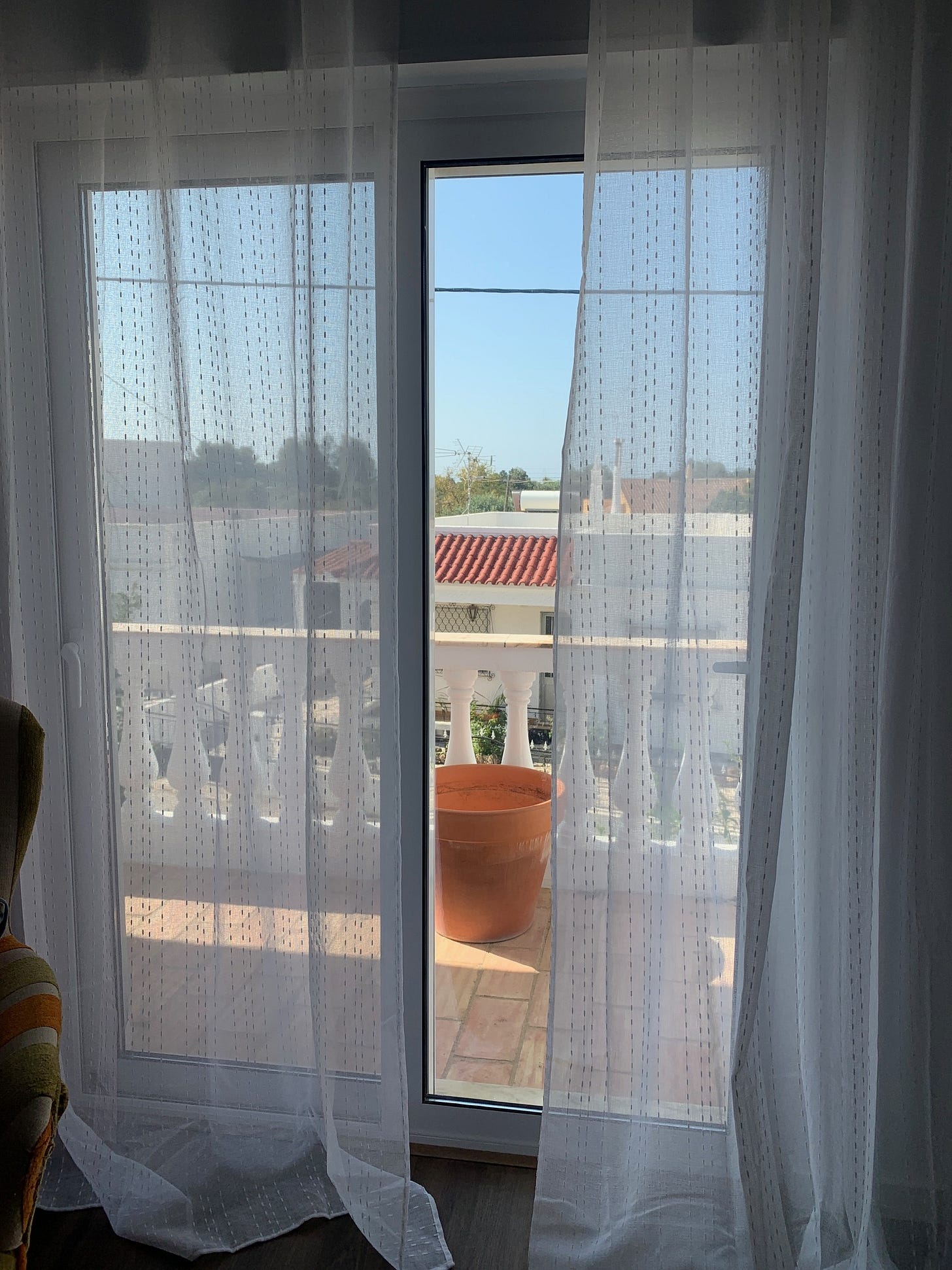 Glass doors out onto a small balcony are flanked by translucent white curtains. Out the door, over the balcony, are red tiled roofs and a bright blue sky.