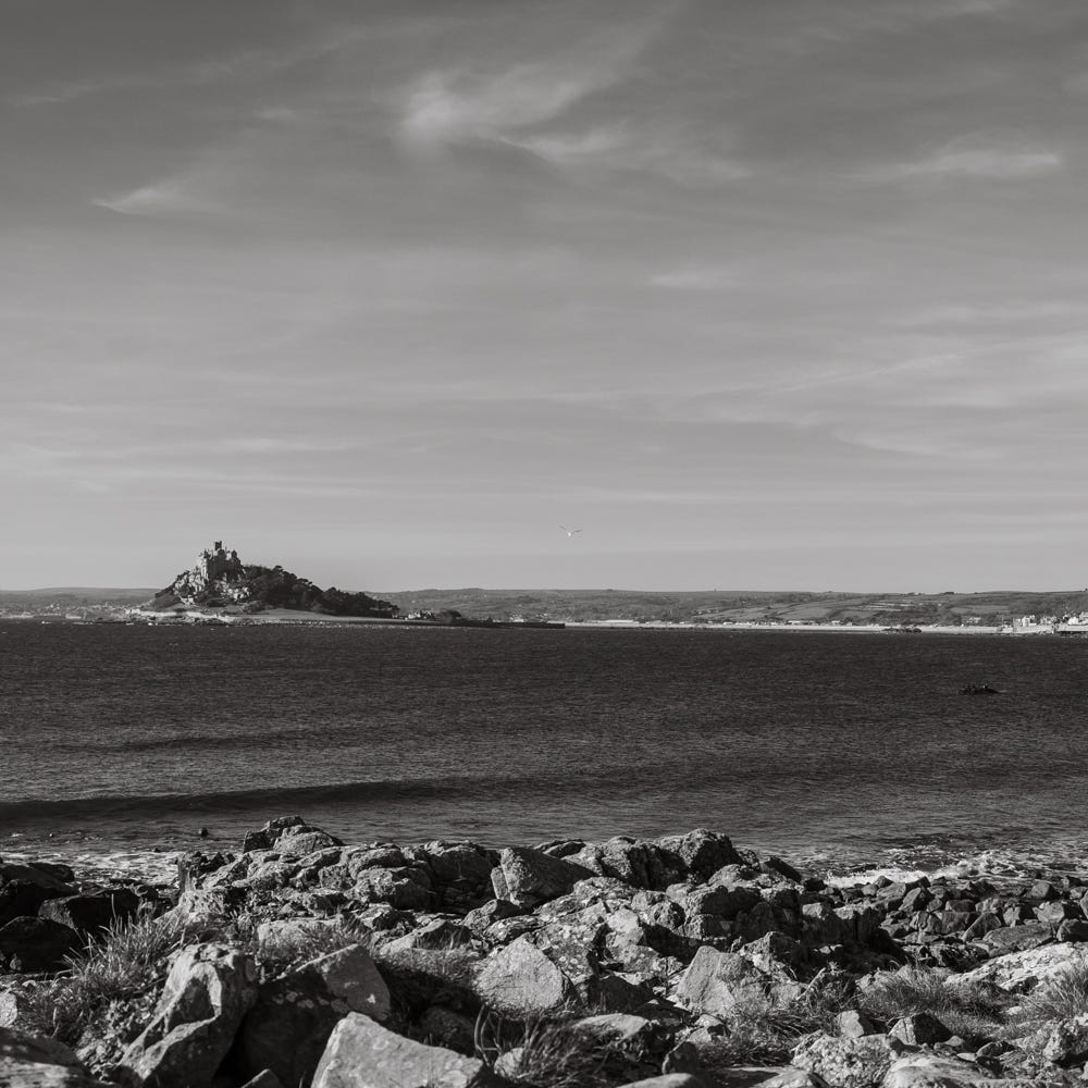 A black-and-white image looking across rocks and the sea to St Michael's Mount, a small island topped by a castle-like building off the coast of Marazion in Cornwall.