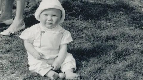 Keir Starmer/Tom Baldwin A baby Keir Starmer sitting on grass, dressed in a bonnet and romper suit