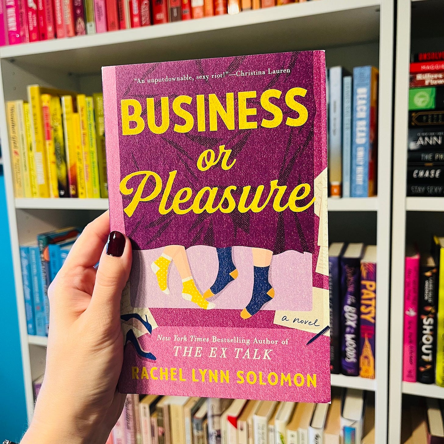 A hand holding the book Business or Pleasure by Rachel Lynn Solomon in front of a bookshelf.