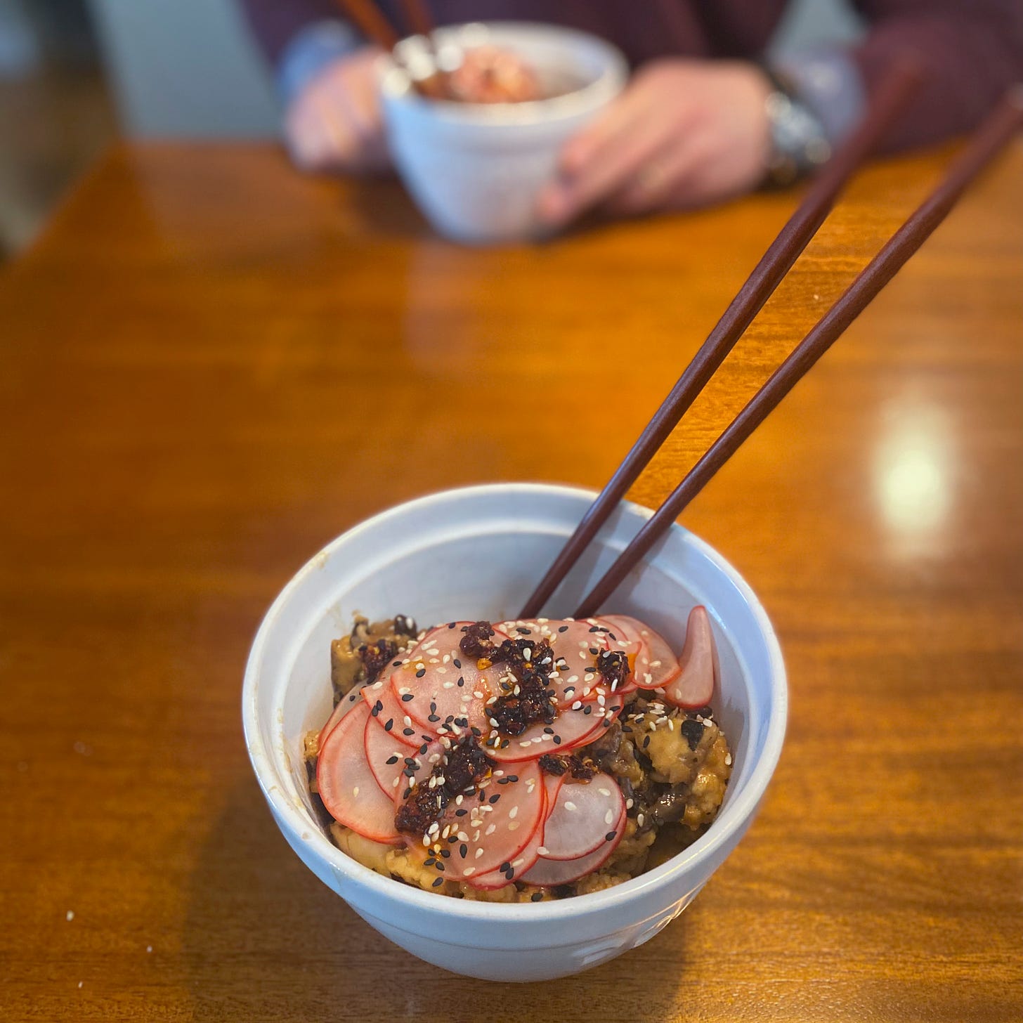 Two white bowls with chopsticks in them across from each other on the table, filled with noodles and tofu and mushrooms in sauce, and topped with thinly sliced pickled radish, sesame seeds, and chili crisp.