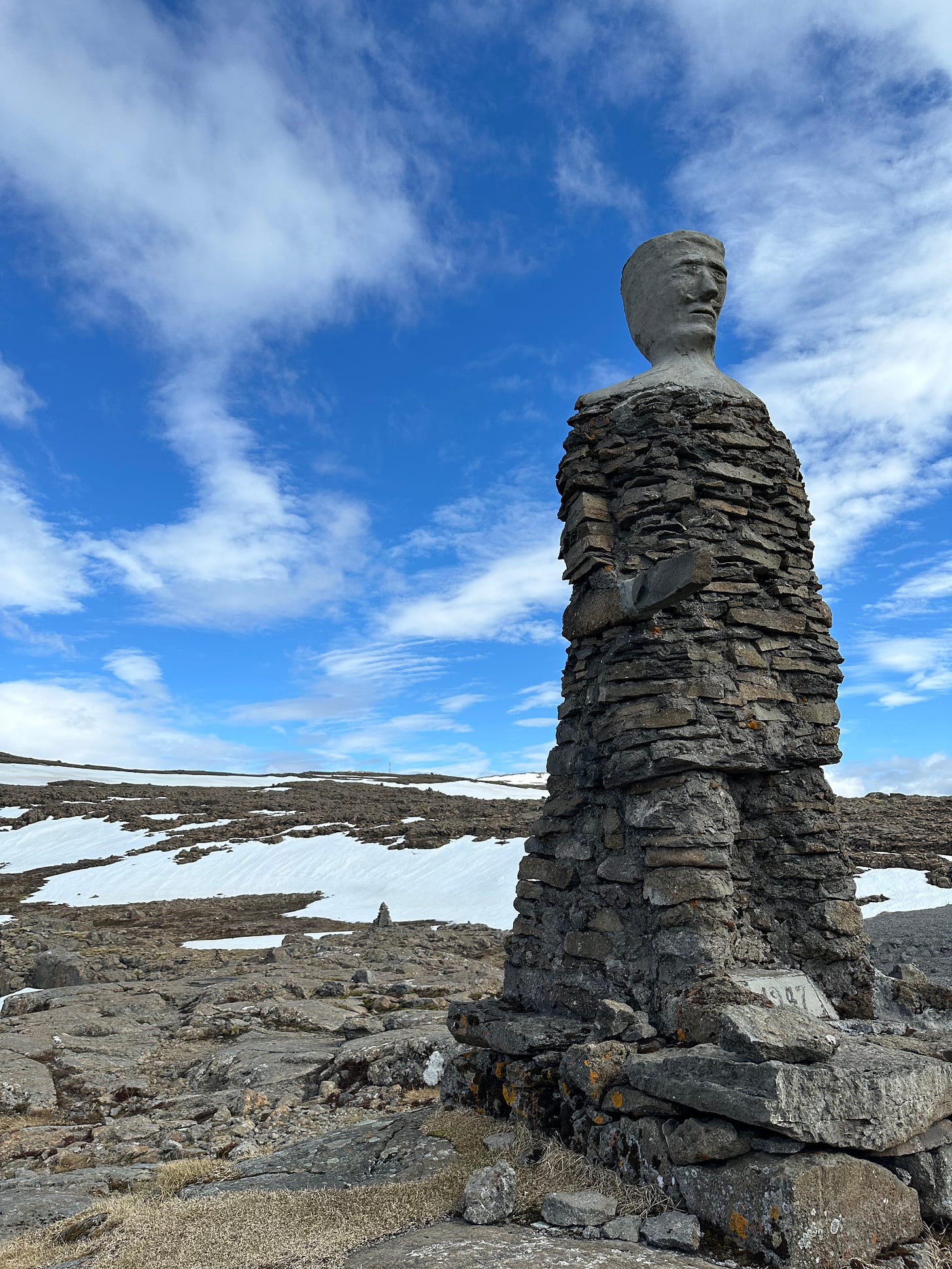 A tall stone cairn shaped like a man with a concrete head and sword pointing the way to Patreksfjordur.  The ground is gray stone with snow patches and the sky is bright blue with small white clouds.