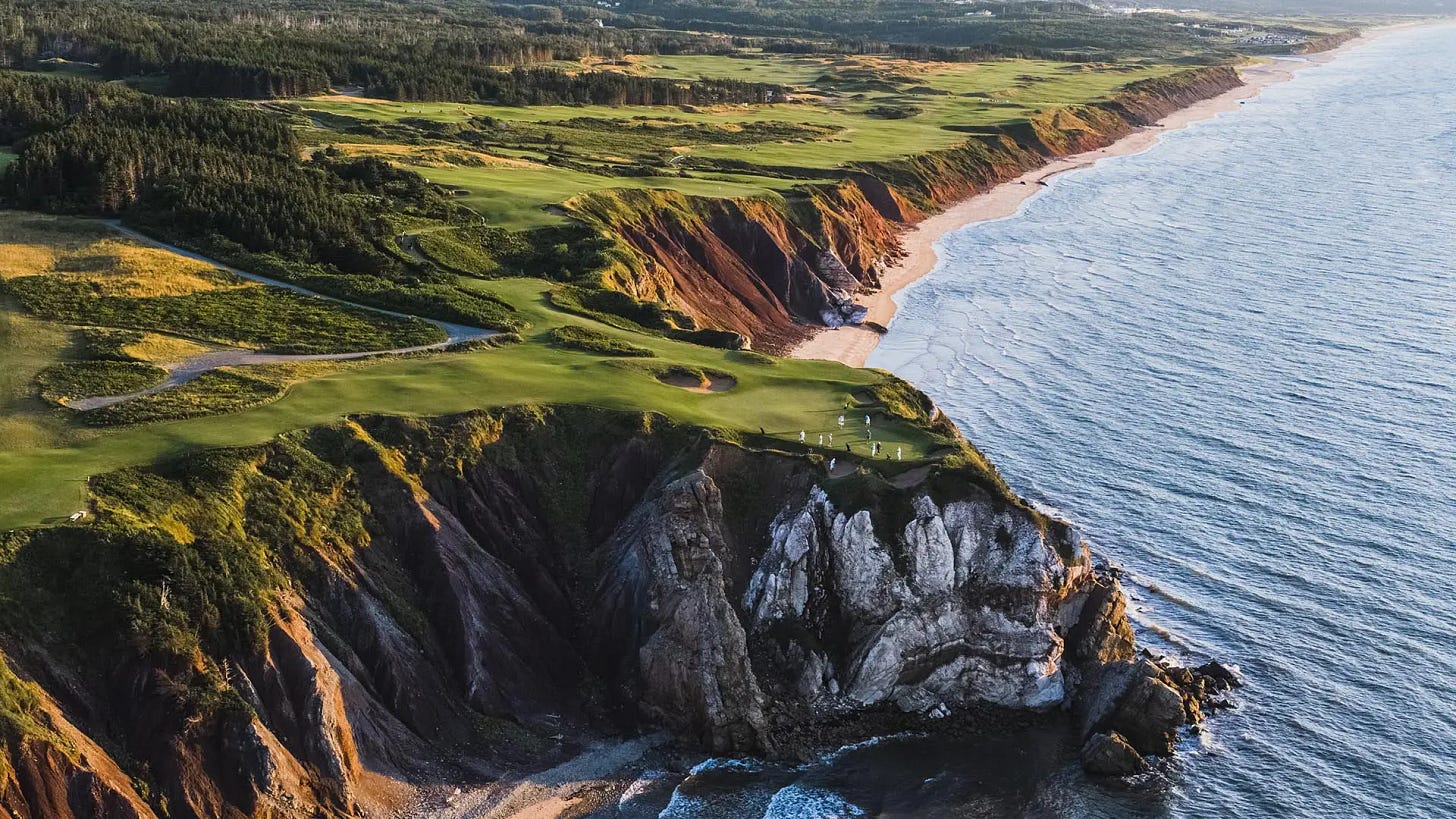 Dramatic aerial view of Cabot Cliffs golf course showing emerald fairways perched atop rugged red cliffs. The coastline stretches into the distance with pine forests inland and the Atlantic Ocean's blue waters meeting the rocky shore. Dramatic aerial view of Cabot Cliffs golf course showing emerald fairways perched atop rugged red cliffs. The coastline stretches into the distance with pine forests inland and the Atlantic Ocean's blue waters meeting the rocky shore.