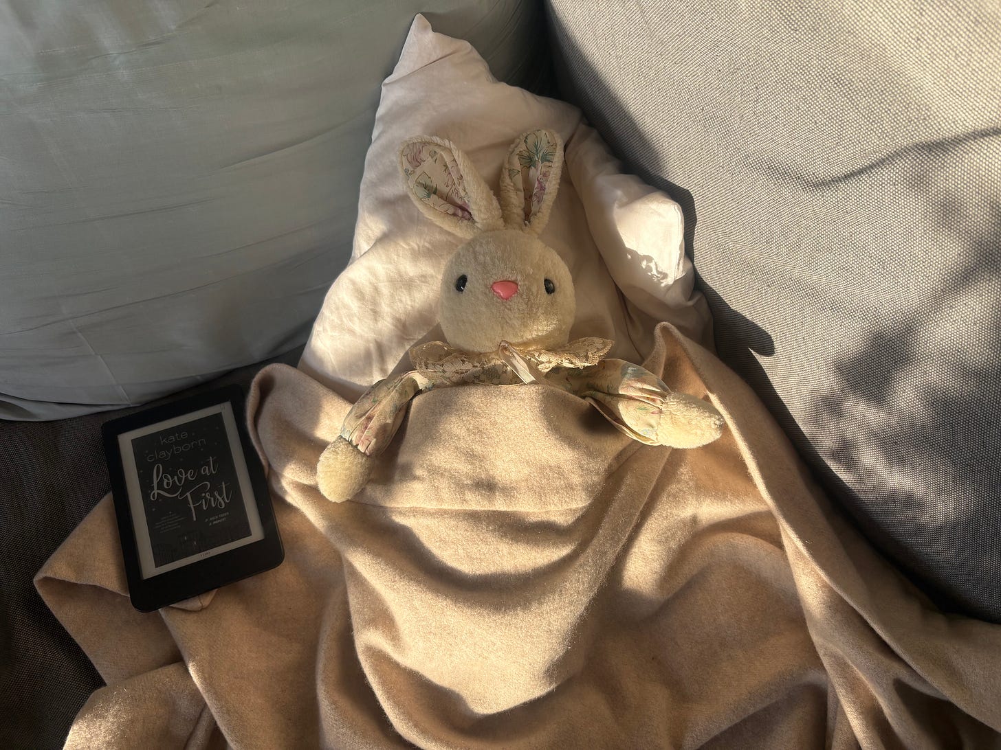 Ein Kuschelhase liegt mit Kuscheldecke auf einem sonnigen Sofa, dameben ein eReader, der das Cover eines Liebesromans (Love at First) anzeigt.