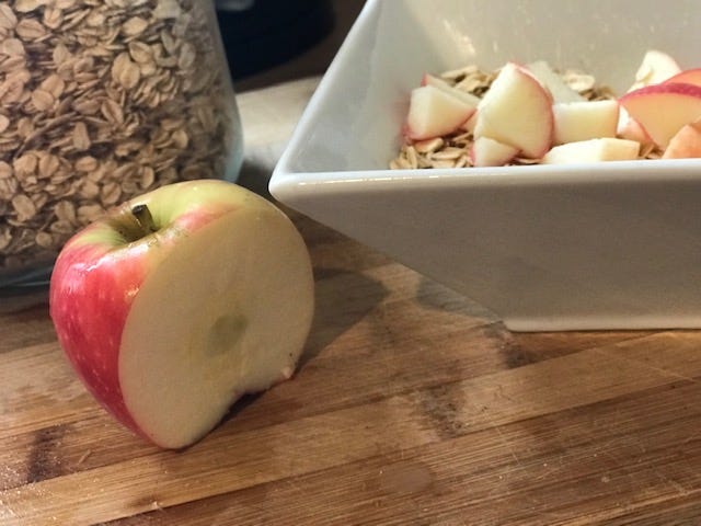 red apple with slice missing and white cereal bowl containing apple pieces next to it with jar of oats in background