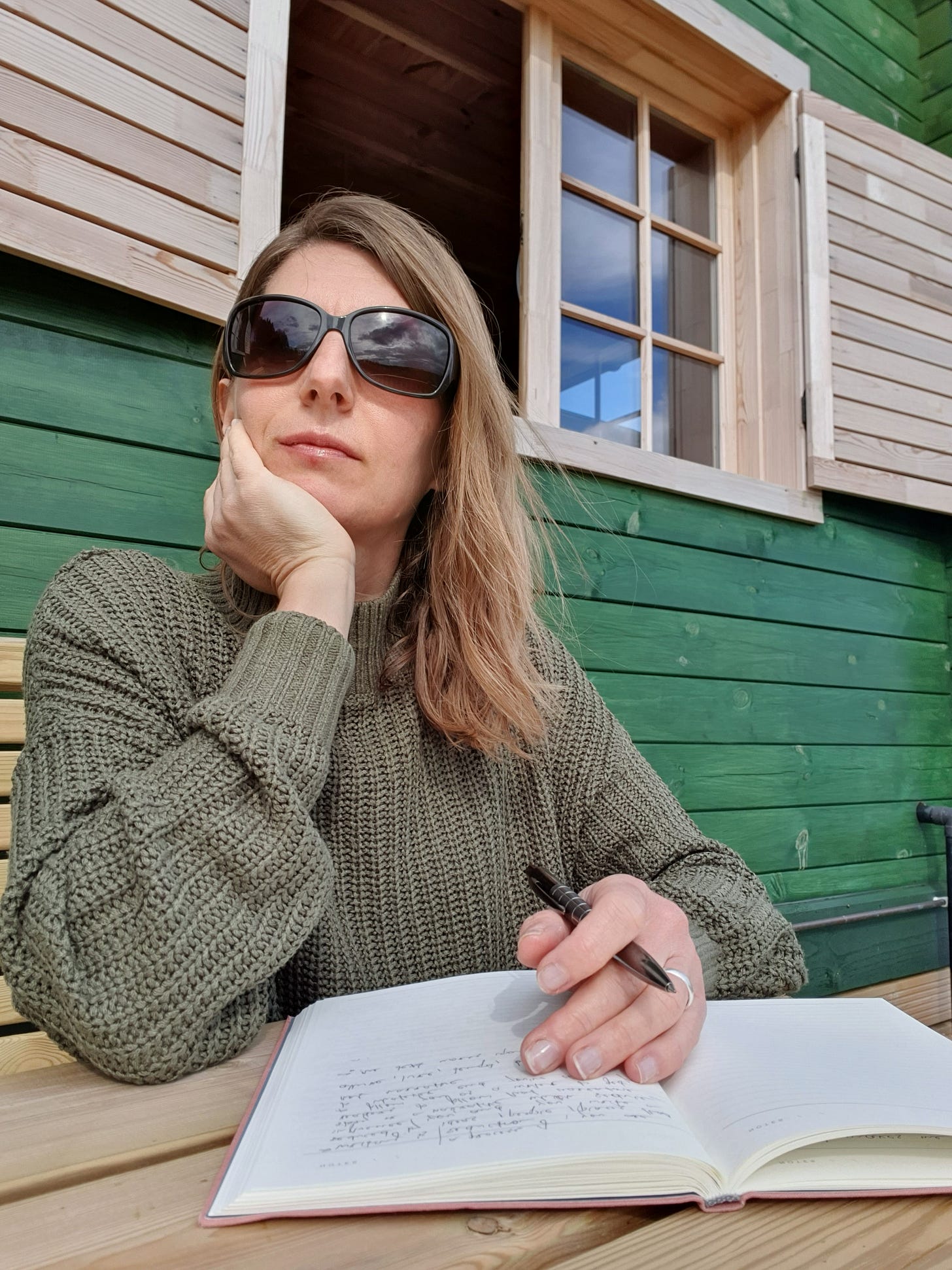 The image shows a woman sitting at a wooden table outdoors. She is wearing a green knitted sweater and large dark sunglasses. Her long, light brown hair is down, and she is resting her chin on her left hand. In her right hand, she is holding a pen over an open notebook with some writing on it. The background features a green wooden wall and a window with white frames. The woman appears to be in a thoughtful or contemplative pose.