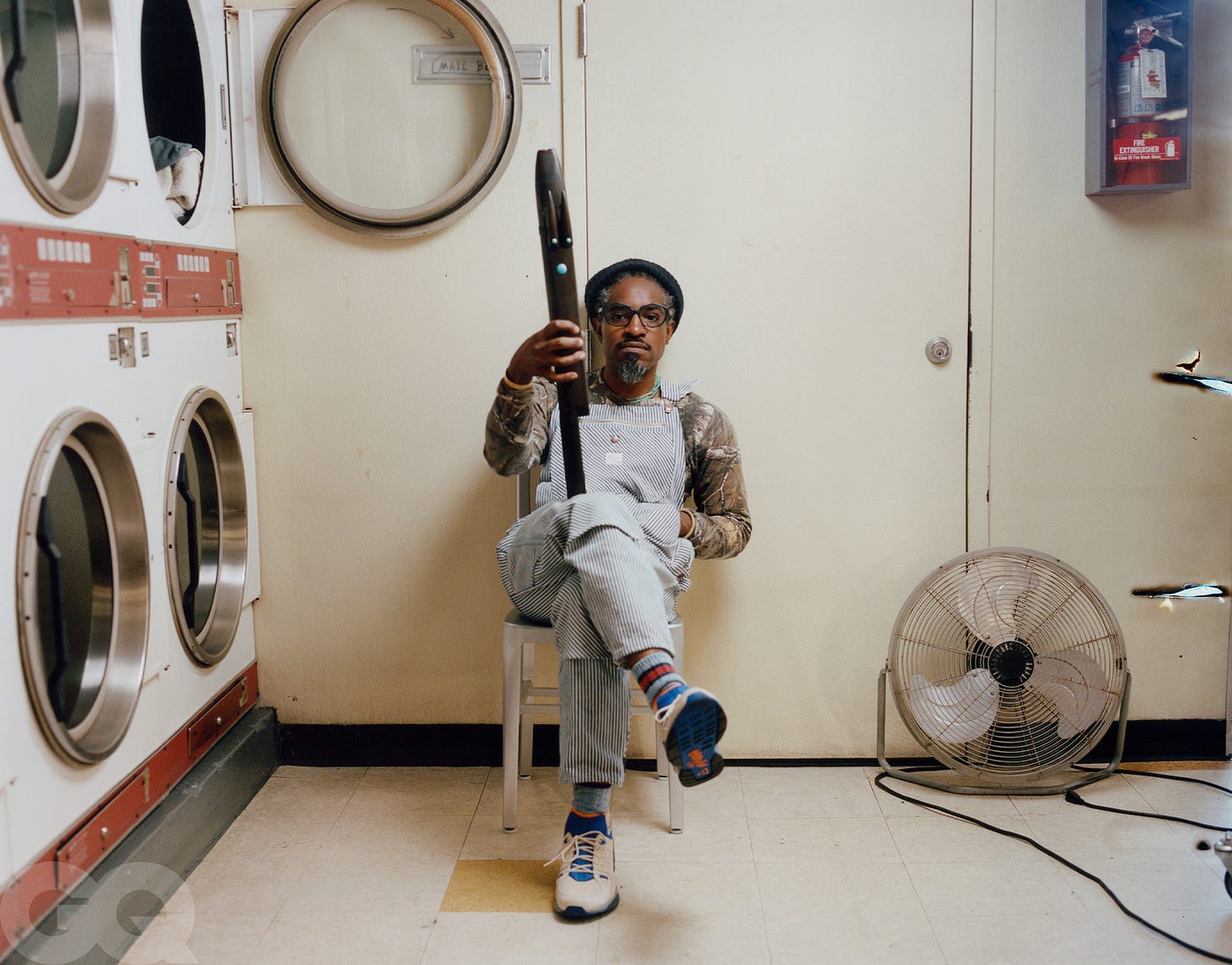 Andre 3000 in striped light blue overalls, with a flue, seated at his laundromat