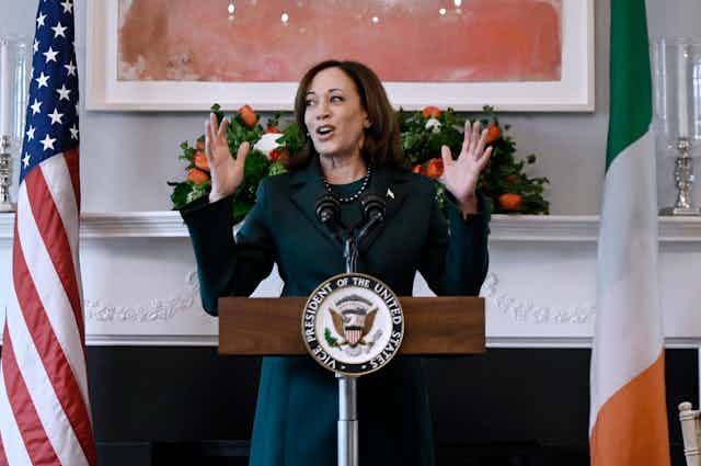 A woman in a green suit stands at a lectern flanked by an American and Irish flag.