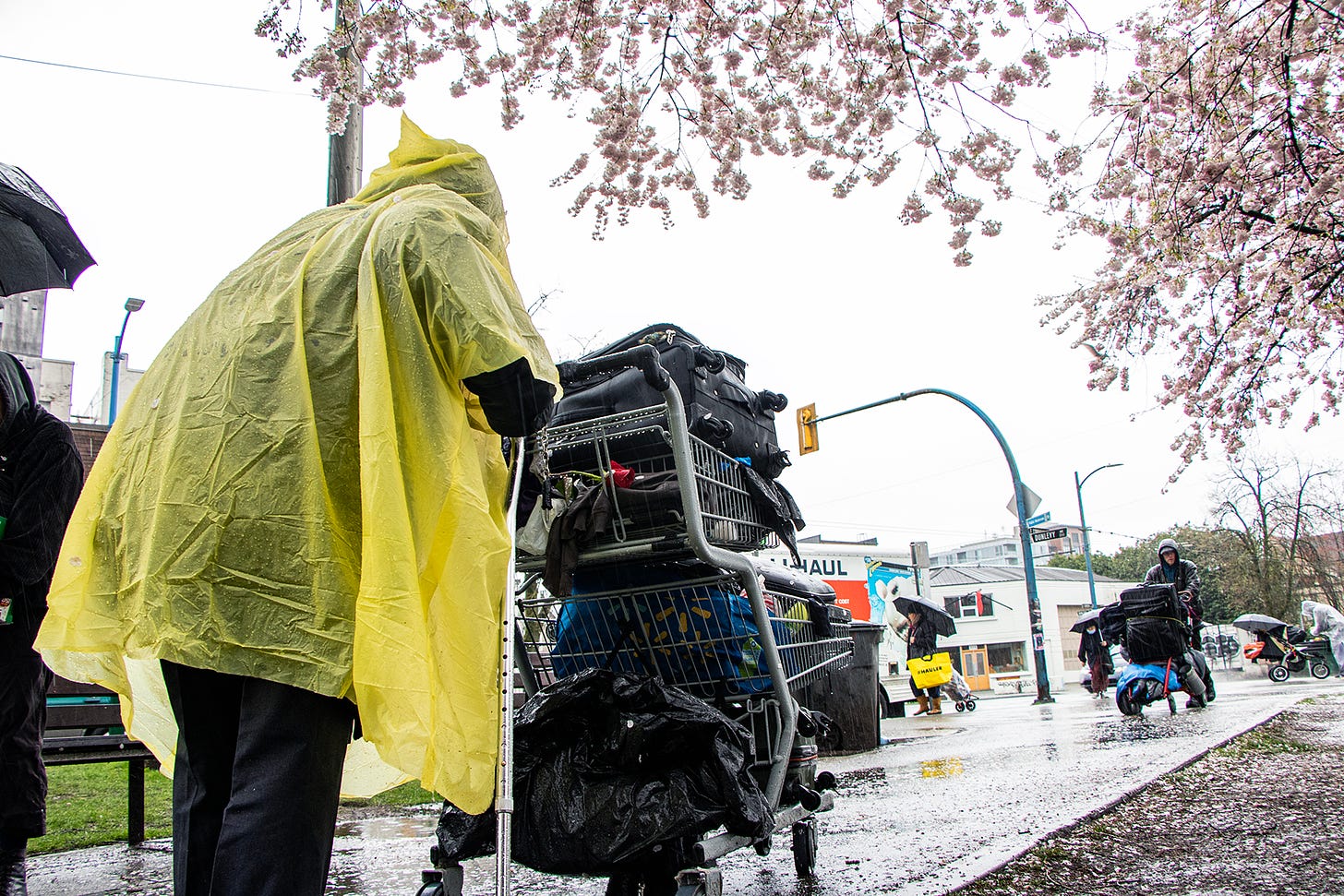 Gurdeep Singh is seen hunched over his shopping cart. In his right hand, he holds a cane, while his left hand supports him by holding onto the shopping cart handle (this is unseen, but implied by his positioning). He's wearing a thin, yellow, plastic rain poncho with his hood up. Above him is a flowering tree. He's on a pathway, and another person is walking towards him with a cart.