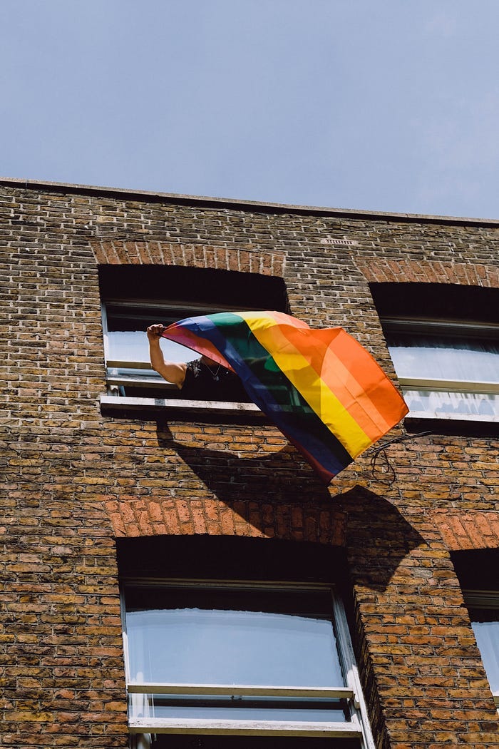 Someone waving a Pride flag through the window of their apartment. Someone waving a Pride flag through the window of their apartment.