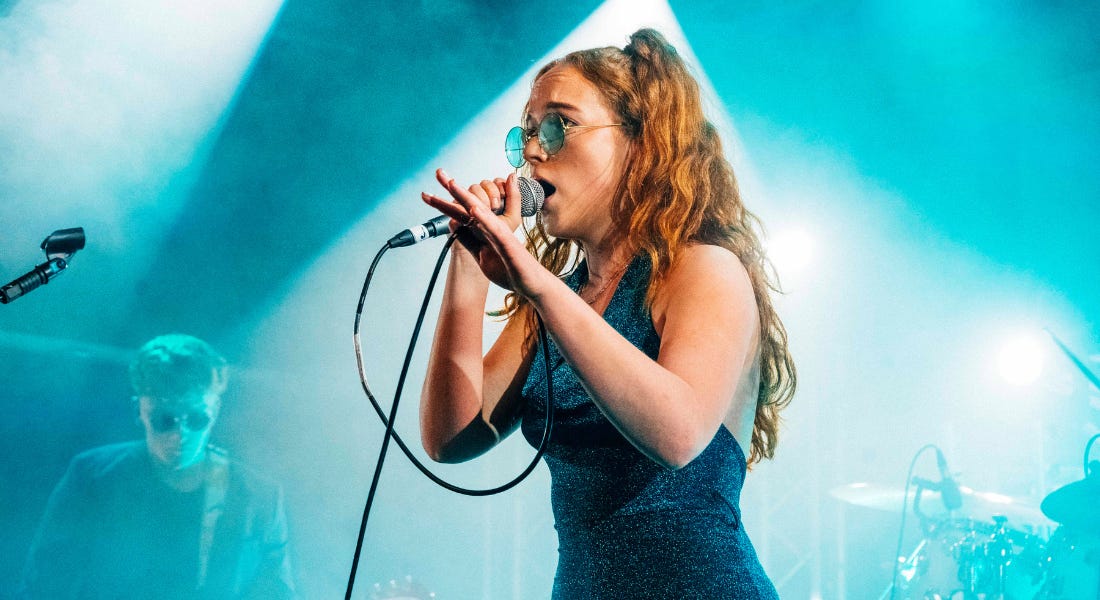 A woman with brown hair wearing blue sunglasses and a blue dress singing into a microphone, with blue lights behind her A woman with brown hair wearing blue sunglasses and a blue dress singing into a microphone, with blue lights behind her
