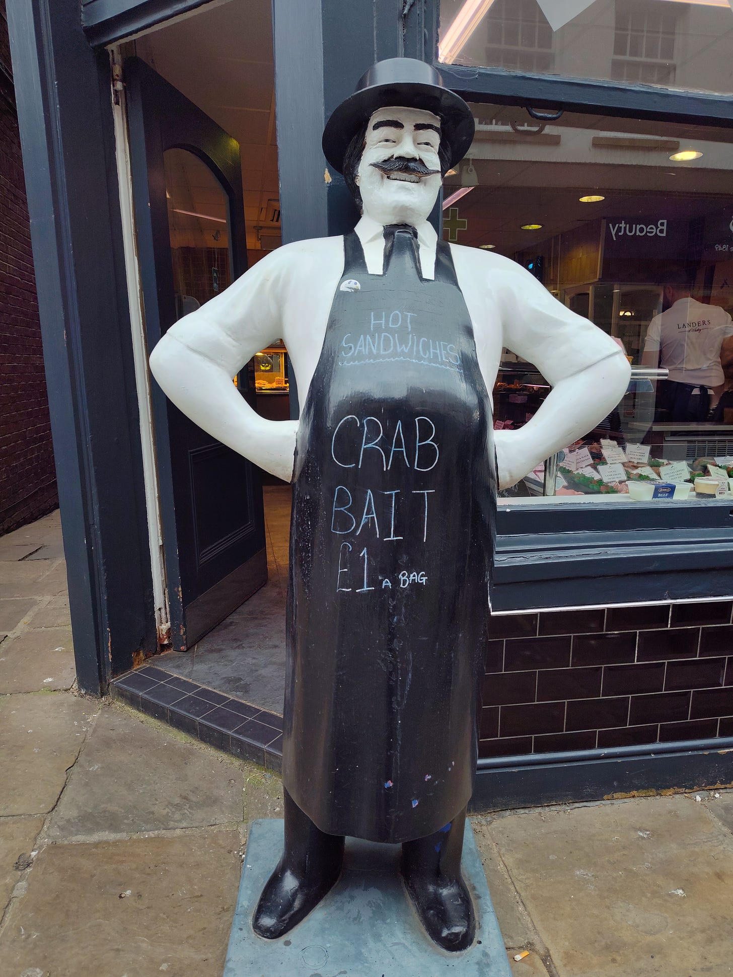 Picture of a statue of a butcher with black mustache, wearing an apron upon which is written HOT SANDWICHES. CRAB BAIT."