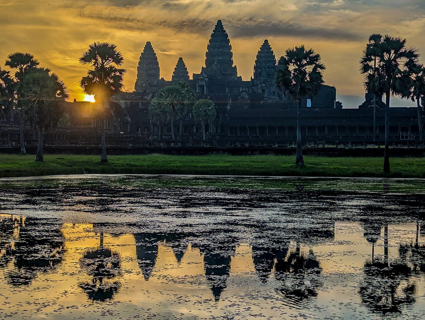 Angkor Wat silhouetted as the sun rises behind it. The sky is yellow and the temple is reflected in the pool outside. 