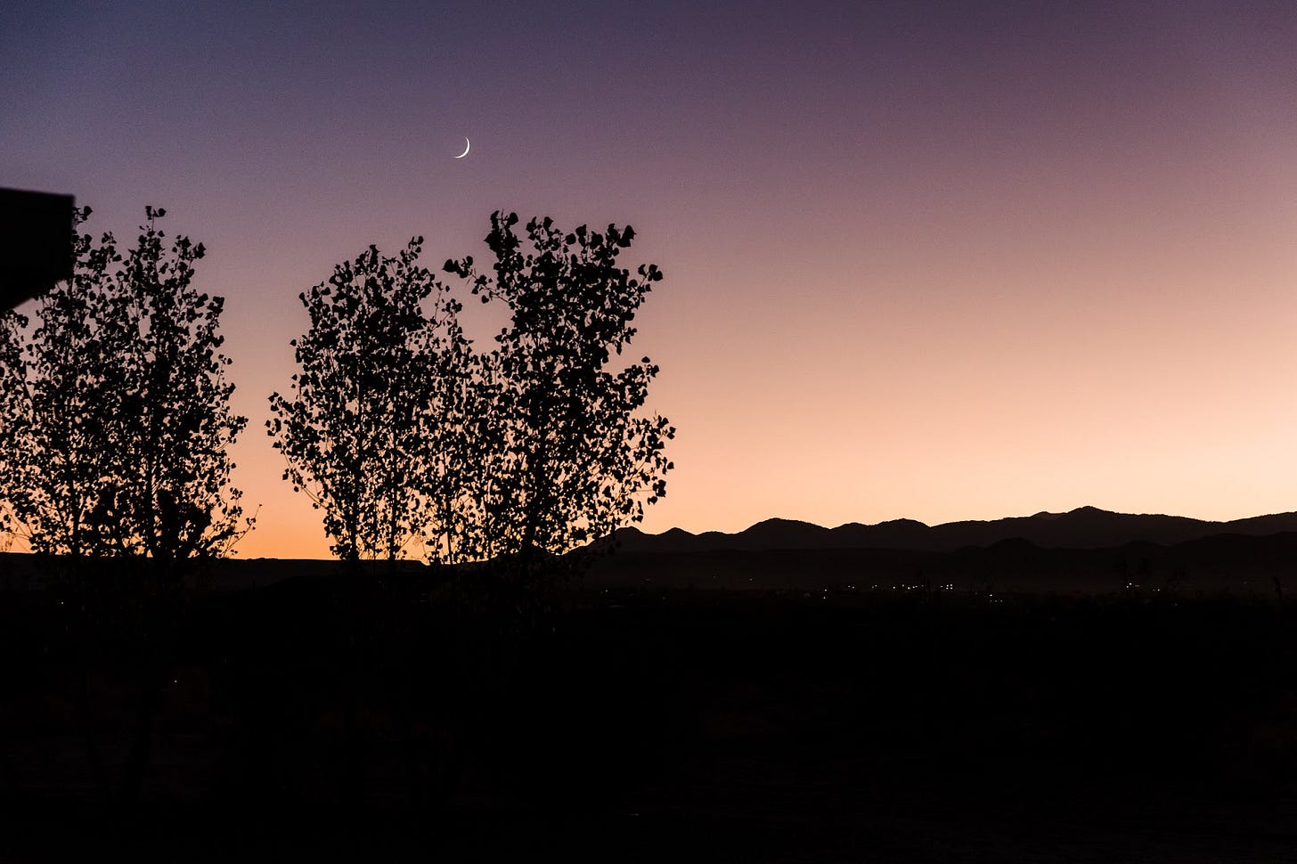 View of the sunset turning to dusk and night, with white, yellow, and orange blending into pink, purple, and blue sky, with a thin, white crescent moon above the outline of pine trees in the foreground and the Little San Bernardino Mountains in the distant background.