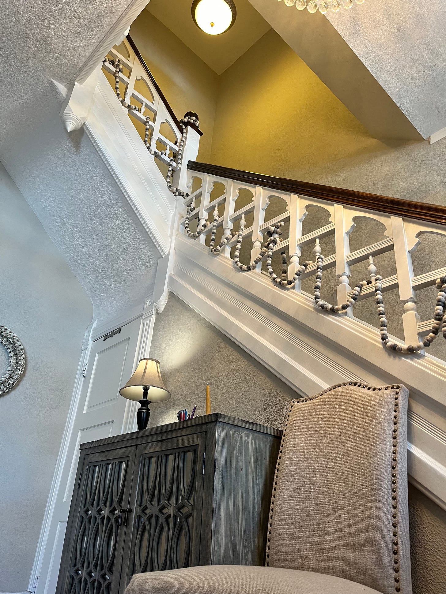 Looking up at a two-story white staircase with brown wood railings and white spindles draped with gray felted wool garland.