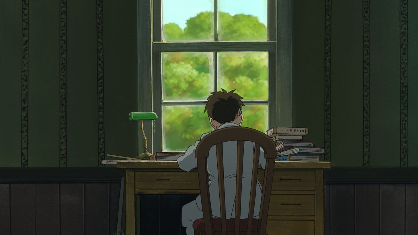 A still from "The Boy and the Heron," with young brunette Mahito sitting at his desk in front of his window, showing the trees outside.