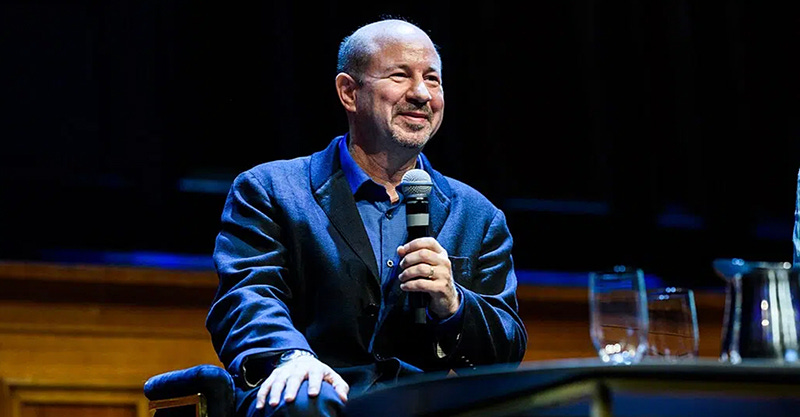 Michael Mann with a microphone on stage. He is light-skinned and balding, wearing a navy-blue suit jacket and shirt