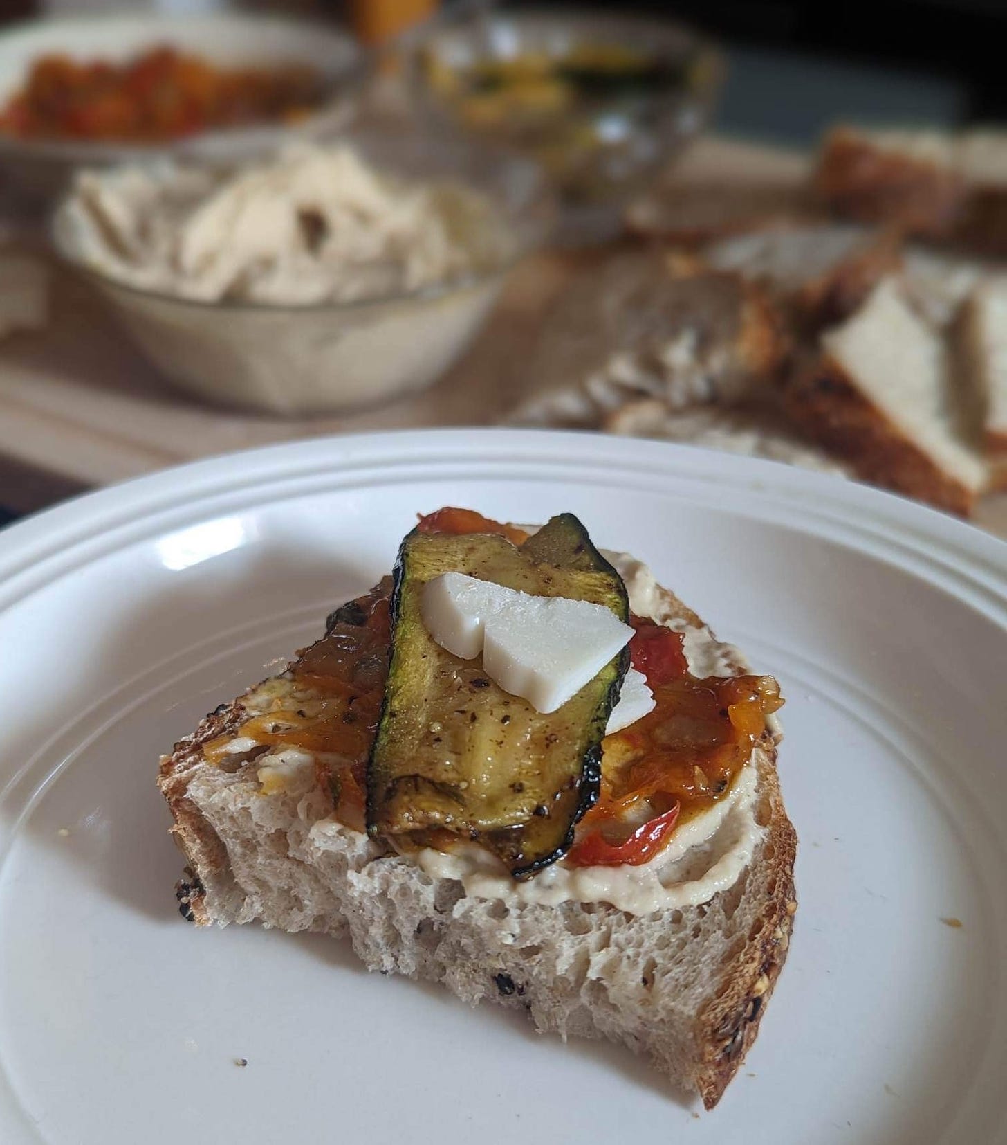 A piece of bread topped with white bean dip, roasted red pepper, grilled zucchini and a vegan Romano cheese. In the background of the image are bowls of dip and slices of bread.