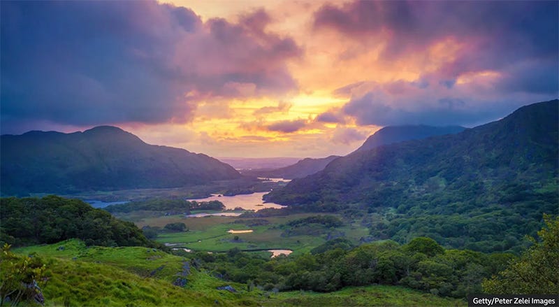 Photo of a sunset with clouds and a lush green landscape with mountains and a river in the background