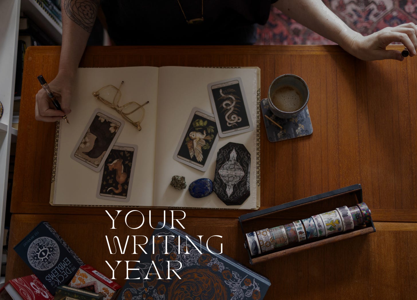 A woman's hands hold a fountain pen and a cap over a notebook covered in tarot cards with a cup of coffee next to it. Text: Your Writing Year