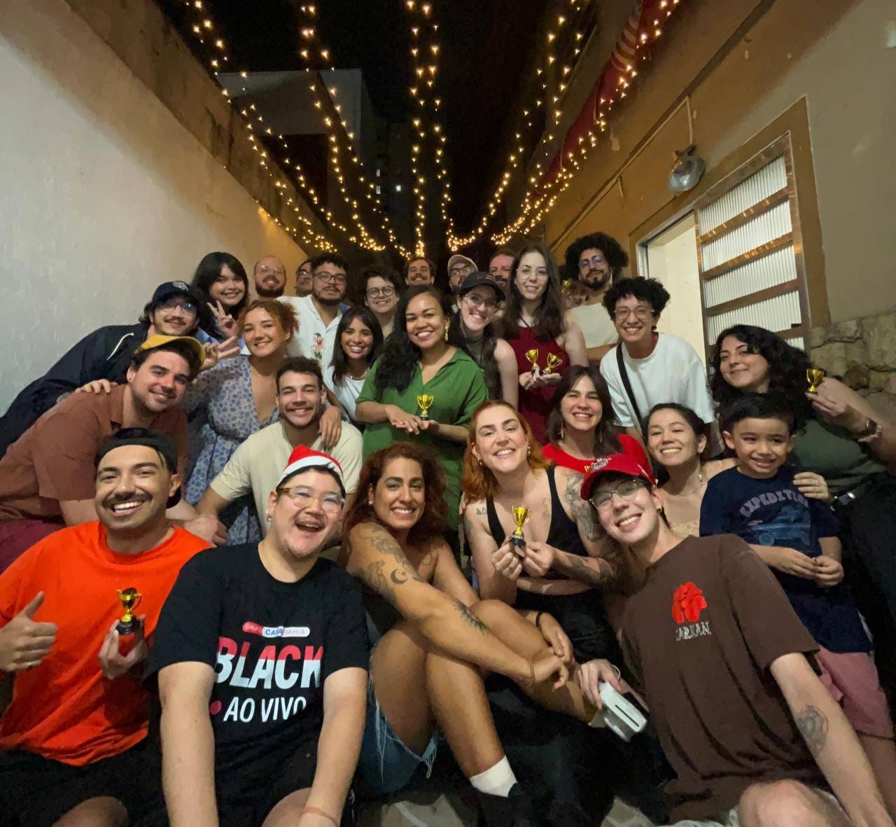 Foto num quintal de noite, com luzinhas de natal penduradas. A foto é de cerca de 27 pessoas, todas ajuntadinhas em fileiras pra caber na foto, sorrindo, algumas com mini troféus nas mãos, com vibe natalina, olhando pra foto.