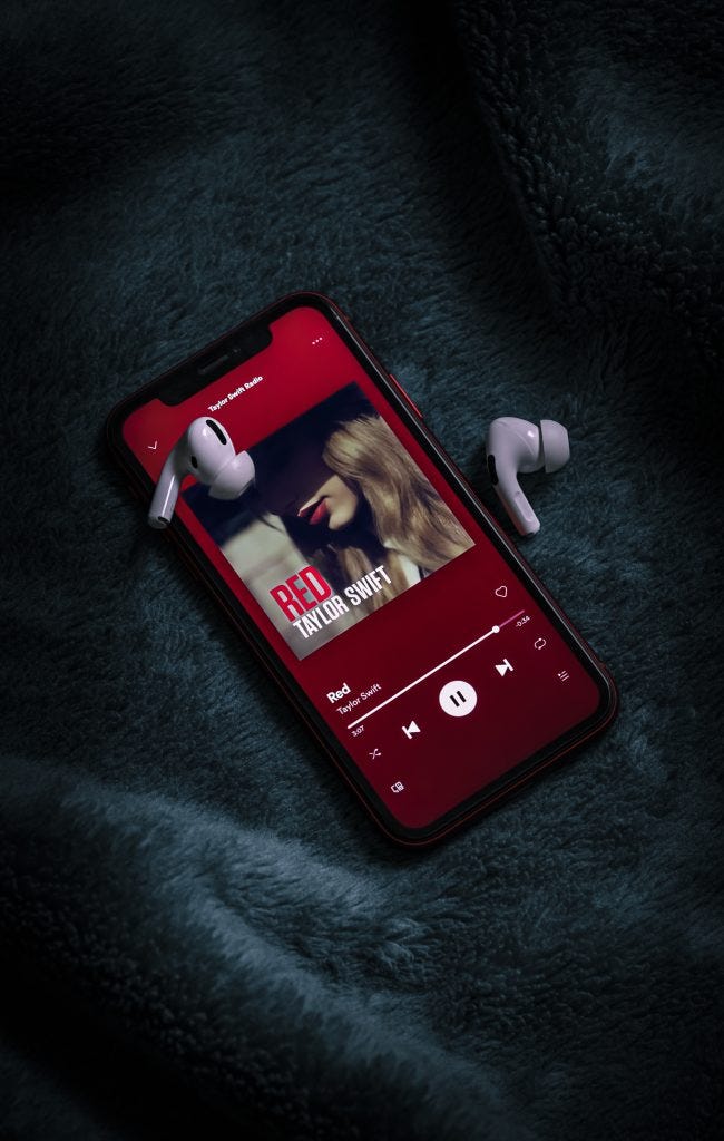 On a navy blanket is a phone and airpods. The phone shows Red by Taylor Swift is playing.
