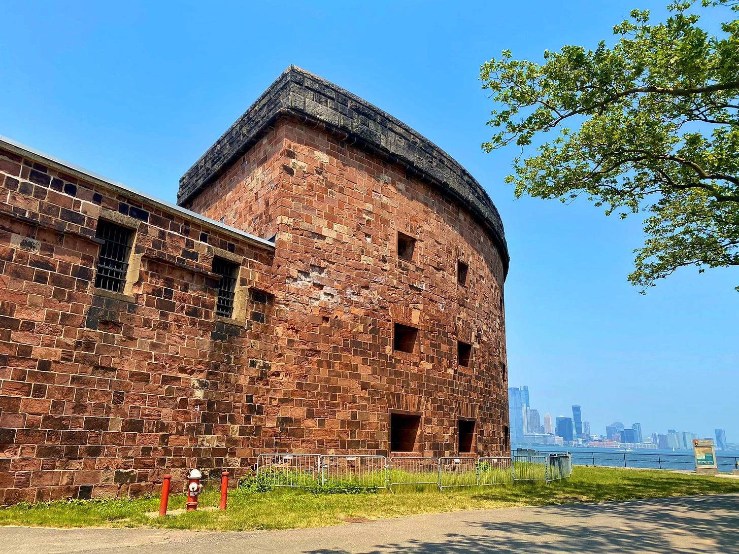 Brick battlements against a blue sky. The Manhattan skyline is visible across the river.