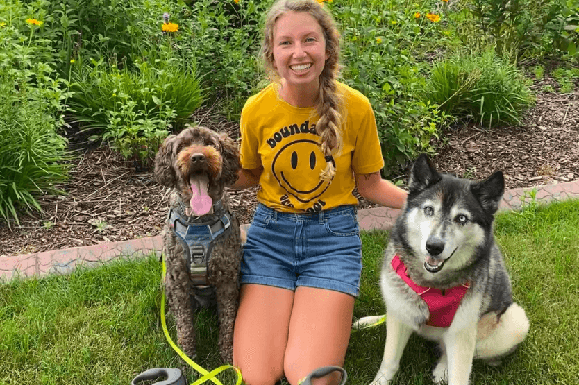 A brown doodle, blonde human in a yellow shirt, and agouti Siberian husky sit next to each other in front of some yellow flowers