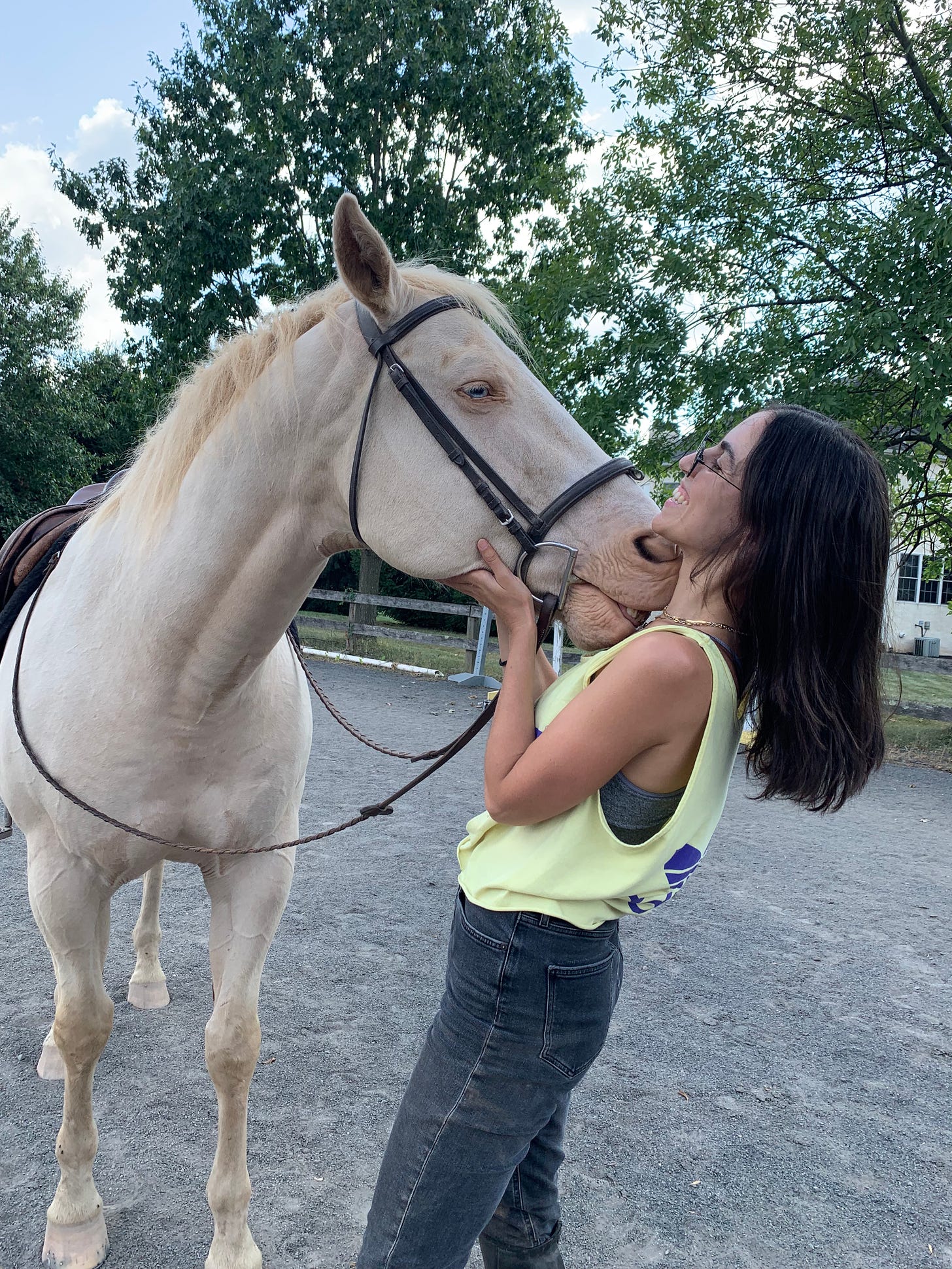 A person with straight dark brown hair and a white horse with blonde hair are nuzzling each other and the person is smiling very wide. The sky is blue and there are trees all around.