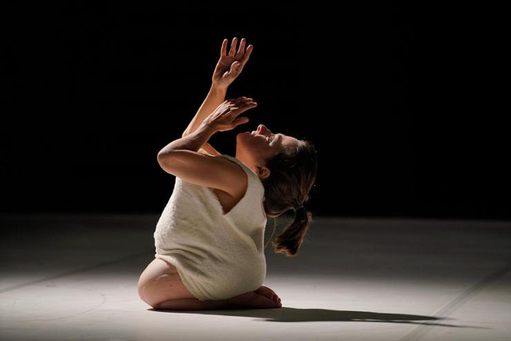 A white femme disabled artist of short stature is performing a work called Gentle Unicorn. She is kneeling, wearing a white dress, with her head drawn back and her hands floating upward toward the sky.