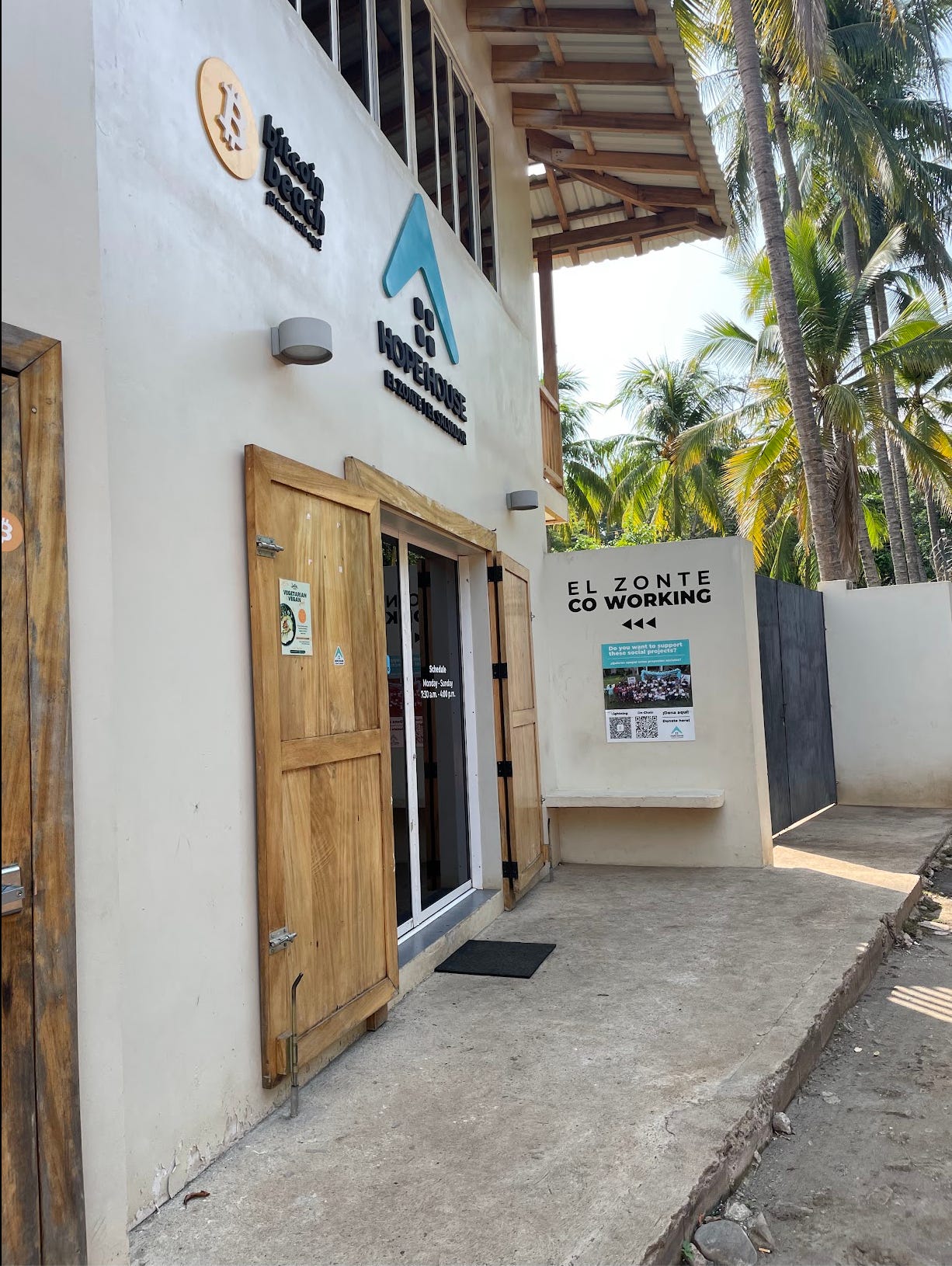 A new building with wooden and glass doors, with numerous signs including one for Bitcoin Beach