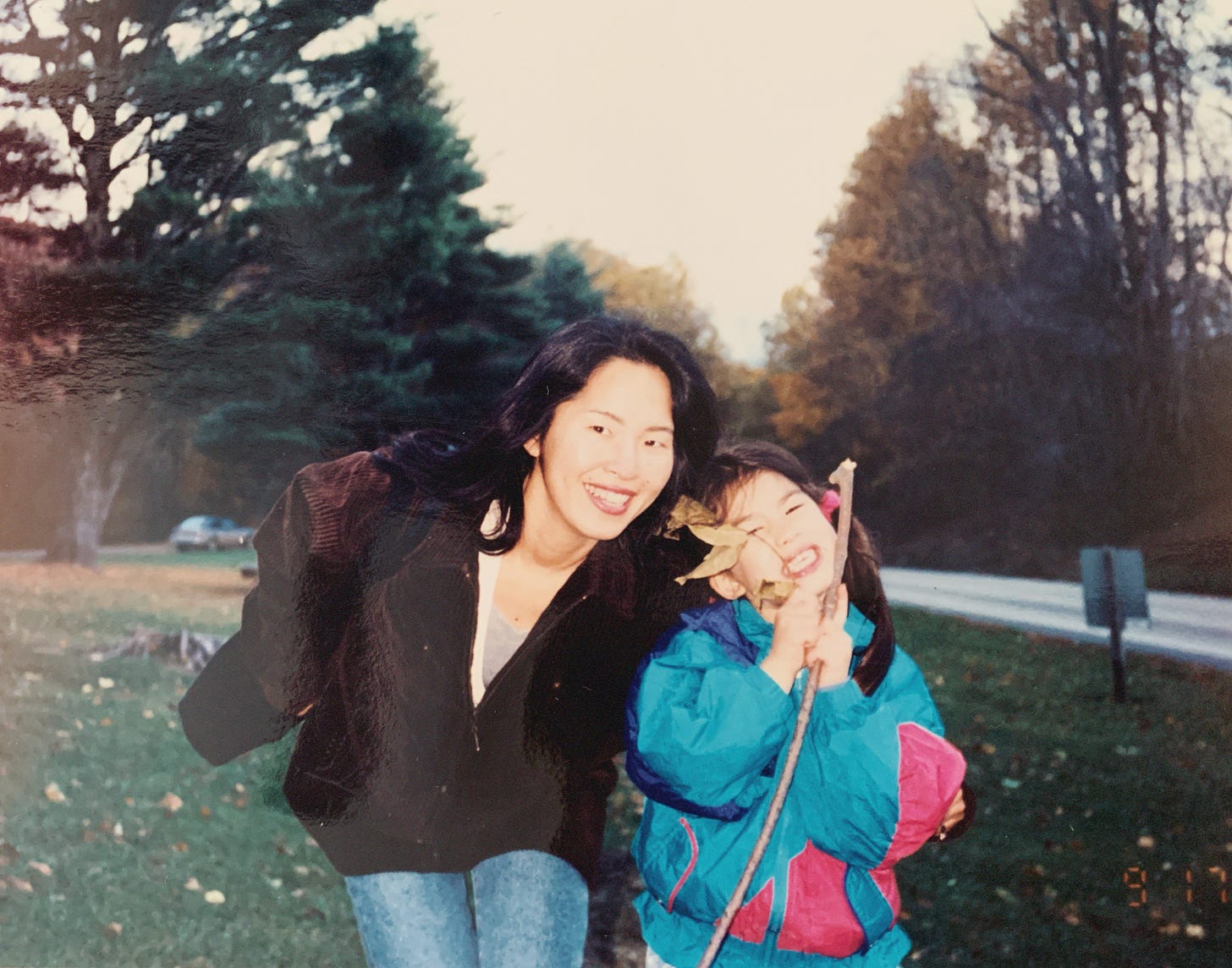 A film photo from the early 90s of the writer's mom, a Japanese woman, with her left arm wrapped around Erika, the writer, at about age four. It's fall, and the trees in the background have begun to change colors. Erika is holding a stick with dried leaves on it. She has a cheesy grin and her mom has almost wind-blown hair with a sweet smile.