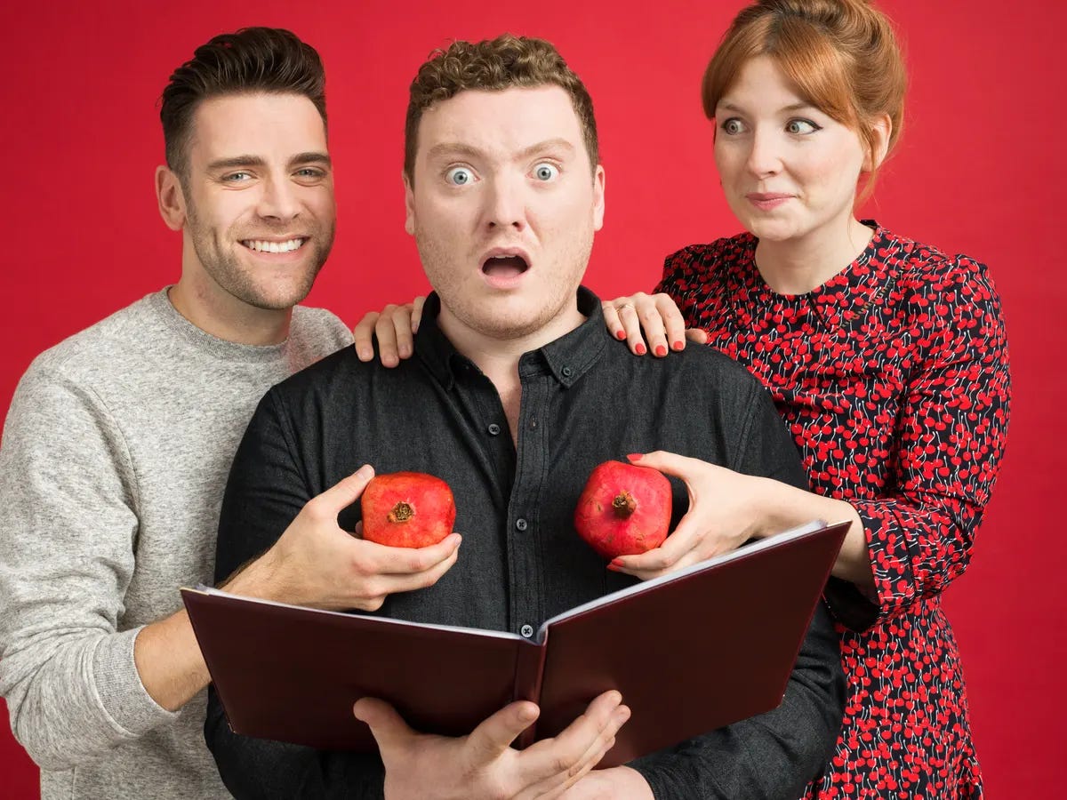 Three white people in casual clothing. A man and a woman hold pomegranates over the chest of another man, who holds a book and looks shocked.