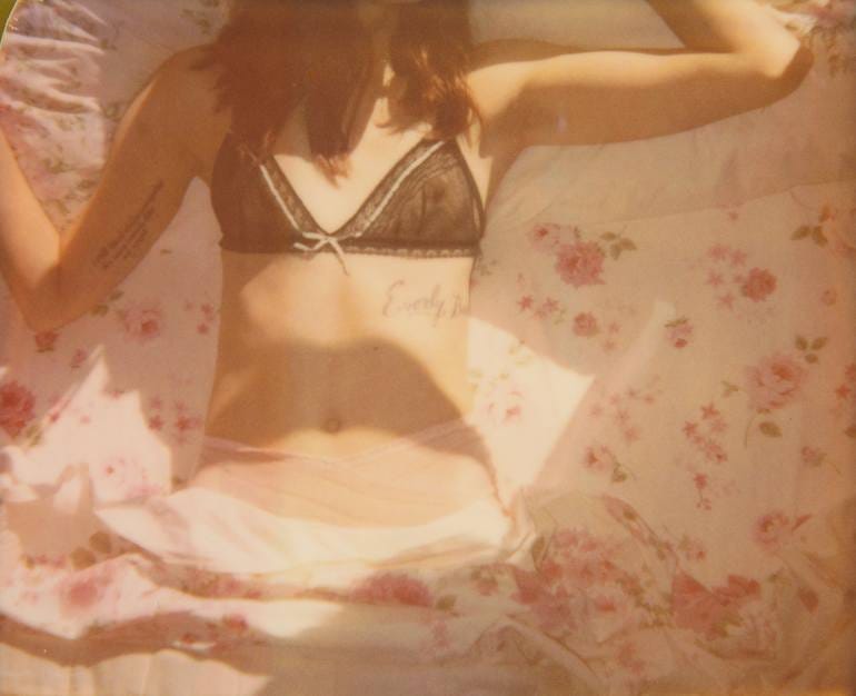 Polaroid: Mulher sem aparecer o rosto, cabelos lisos até os peitos, usando sutiã transparente verde, hot pant rosa deitada sobre um lençol florido