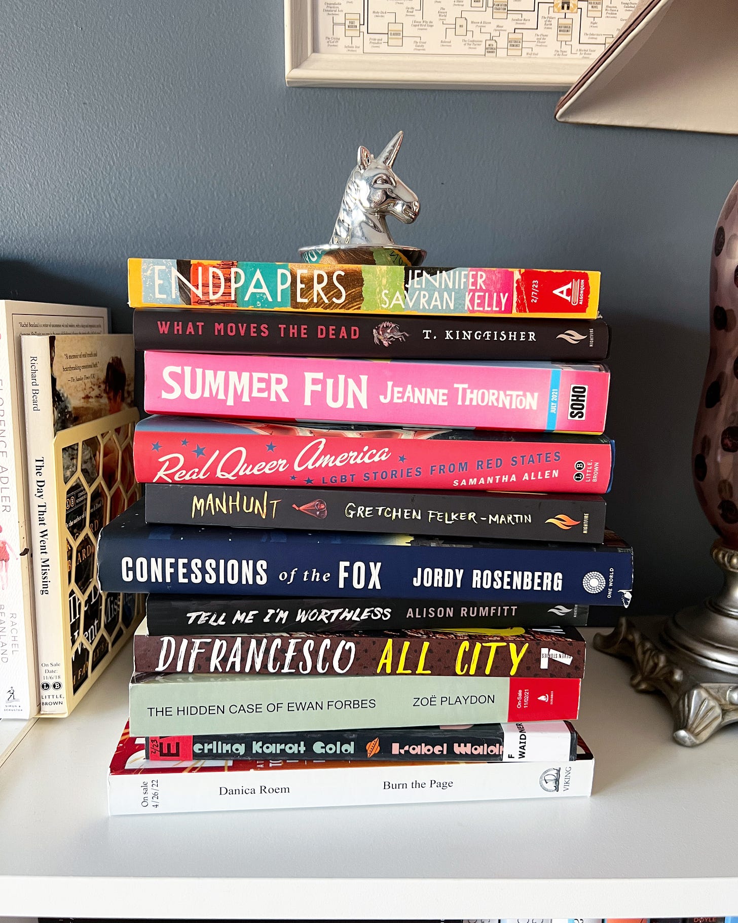 A stack of trans books with a silver unicorn head on top. The books include Endpapers, What Moves the Dead, Summer Fun, Real Queer America, Manhunt, Confessions of the Fox, Tell Me I'm Worthless, All City, The Hidden Case of Ewan Forbes, and Burn the Page. 