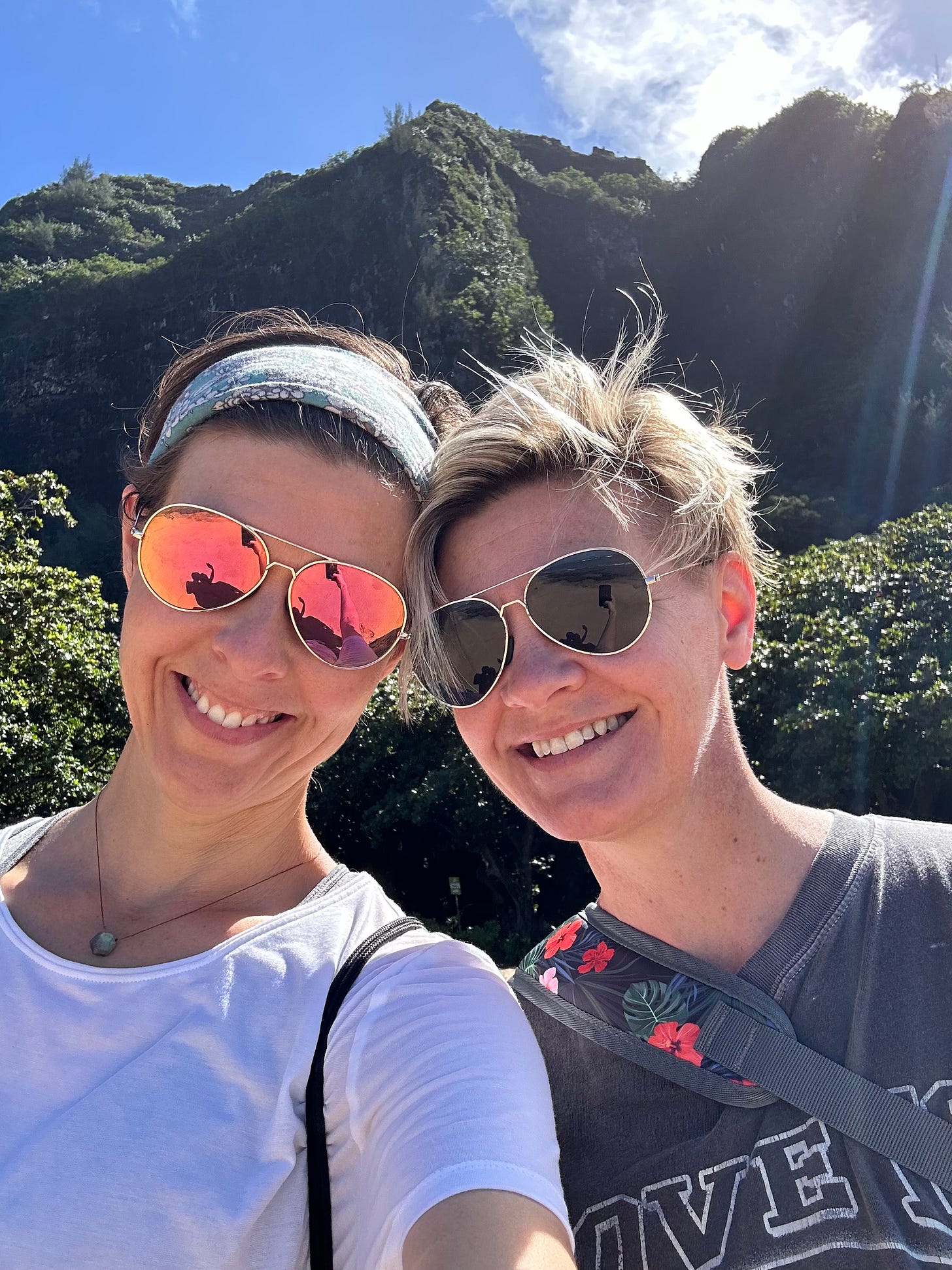 Two women with short hair and sunglasses smile in front of the green mountains and blue sky of Kauai.